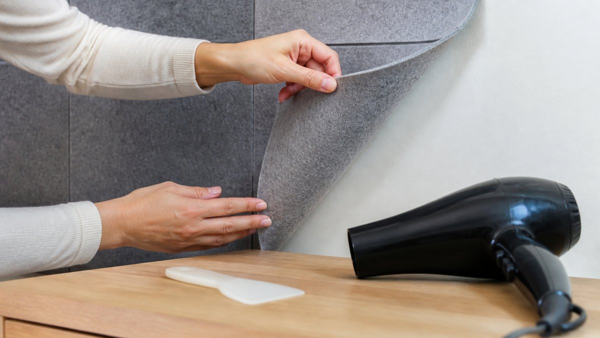 Will Adhesive Wall Panels Damage Paint? What to Know 2 Close-up of hands gently lifting a corner of a fabric wall panel away from a painted wall, with a hairdryer nearby and a plastic scraper resting on a table